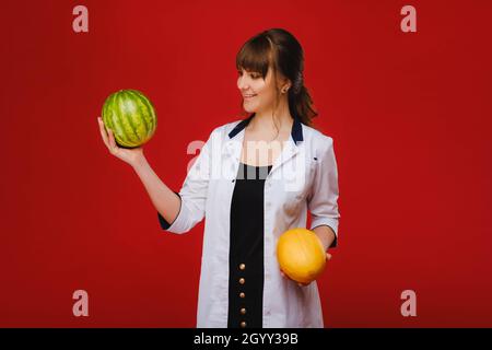 una dottore femminile in un cappotto bianco con frutta nelle mani si pone su sfondo rosso, melone, anguria Foto Stock