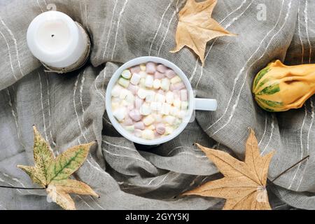Tazza di cacao con marshmallows su sfondo tessile grigio con foglie autunnali, zucca e candela. Vista dall'alto, piatto. Foto Stock