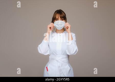 Una ragazza medico si trova in una maschera medica, isolato su uno sfondo grigio Foto Stock