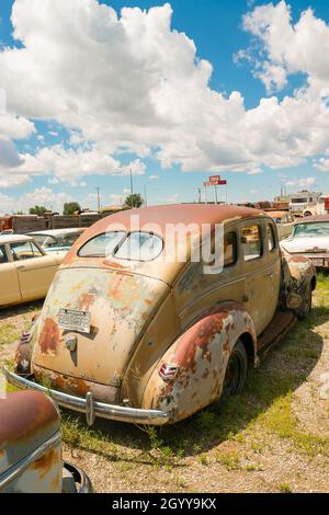 Un'auto ford ford deluxe d'epoca arrugginita parcheggiata su un terreno erboso circondata da altre auto d'epoca sotto un cielo parzialmente nuvoloso in un deposito di rottami. Foto Stock