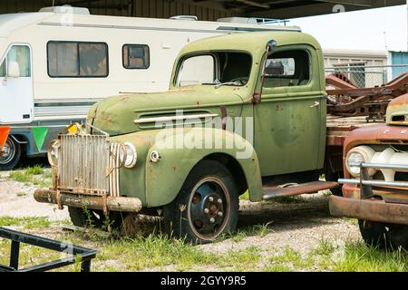 Un pick-up vintage verde con macchie di ruggine si trova in un cortile accanto a un vecchio camper e a un'auto d'epoca. Foto Stock