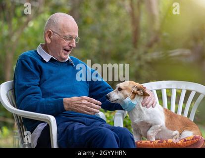 Uomo anziano seduto in giardino e ridendo al cane che indossa maschera protettiva Foto Stock