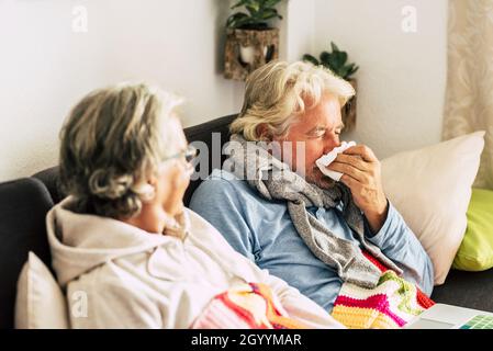 Vecchia donna con suo marito starnutire in carta tissue a casa. Coppia anziana rilassarsi sul divano in malattia a causa del virus. Coppia anziana in abiti caldi Foto Stock