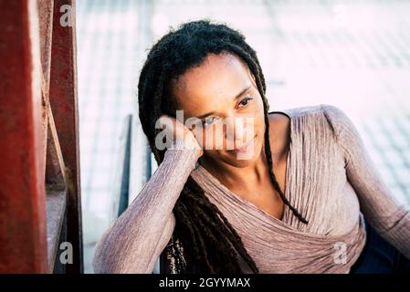 Donna afroamericana pensierosa sorridente con testa in mano. Donna nera con capelli deadlock. Giovane donna seduta sul banco del parco. Elegante Wi donna Foto Stock