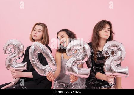 Un gruppo di girlfriends su sfondo rosa con palloncini in lamina d'argento nella forma dei numeri 2022 stanno sperimentando emozioni diverse, celebrando Foto Stock
