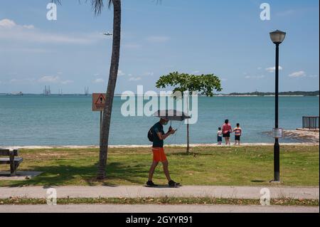 09.10.2021, Singapore, Repubblica di Singapore, Asia - l'uomo che detiene un ombrello cammina lungo la riva al Changi Beach Park mentre un aereo si avvicina. Foto Stock