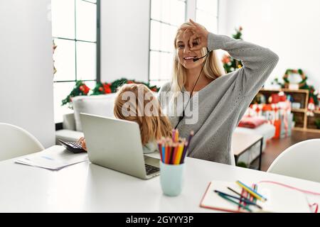 Madre della figlia caucasica che indossa la cuffia dell'agente del call center a casa sorridendo felice di fare ok segno con la mano sull'occhio guardando attraverso le dita Foto Stock