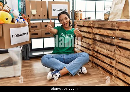 Giovane donna afroamericana che indossa la t shirt del volontario alle donazioni stand approvando fare il gesto positivo con la mano, i pollici in su sorridendo e felice per Foto Stock