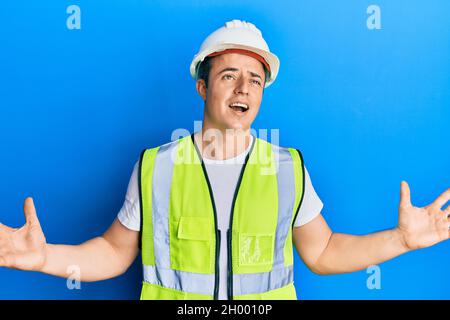 Bel giovane uomo che indossa casco di sicurezza e giacca riflettente pazzo e pazzo urlando e urlando con espressione aggressiva e braccia sollevate. Frustrazione Foto Stock