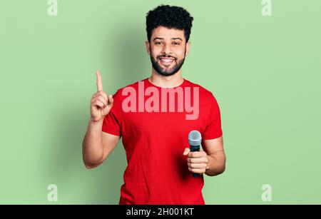 Giovane uomo arabo con la barba cantare canzone utilizzando il microfono sorridendo con un'idea o domanda puntando dito con volto felice, numero uno Foto Stock