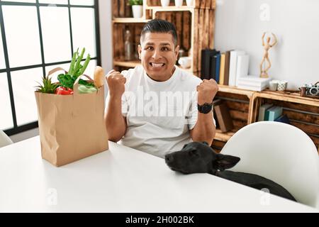 Giovane ispanico seduto con sacchetto di carta con generi alimentari urlando orgoglioso, celebrando la vittoria e il successo molto eccitato con le braccia sollevate Foto Stock