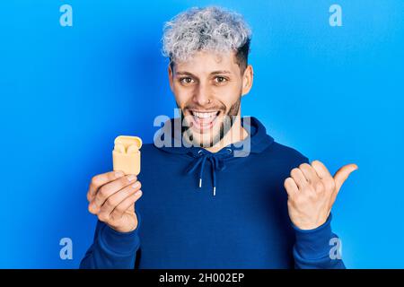 Giovane uomo ispanico con moderni capelli tinti tenendo auricolari senza fili puntando il pollice verso il lato sorridendo felice con bocca aperta Foto Stock