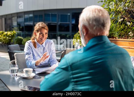 Giovane donna graziosa di affari e uomo maturo che firma l'accordo dentro caffetteria all'aperto Foto Stock