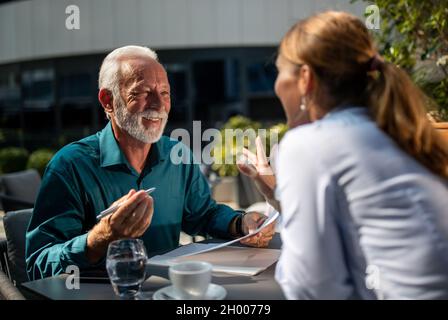 Giovane donna graziosa di affari e uomo maturo che firma l'accordo dentro caffetteria all'aperto Foto Stock