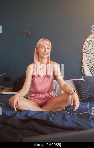 Modello femminile con lunghi capelli rosa in eleganti pigiami, in posa di loto su un comodo letto Foto Stock
