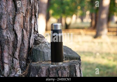 Thermos a vuoto neri su un ceppo di albero. Sfondo della foresta sfocato, spazio di copia. Foto Stock