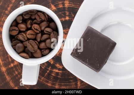 Una tazza di chicchi di caffè e una caramella al cioccolato, in primo piano, con un piattino bianco su una pietra ardesia, vista dall'alto in primo piano. Foto Stock