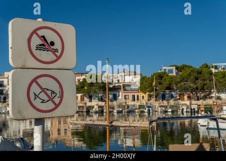 Primo piano di un cartello che vieta il bagno e la pesca nella città di Portocolom. Sullo sfondo, fuori fuoco, il molo con le barche ormeggiate e riflettere Foto Stock