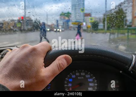 vista della mano del conducente sul volante della vettura sullo sfondo dell'incrocio durante la pioggia Foto Stock