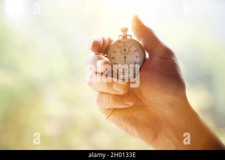 La mano di un uomo tiene un classico cronometro meccanico analogico in metallo cromato, su uno sfondo verde sfocato. Foto di alta qualità Foto Stock