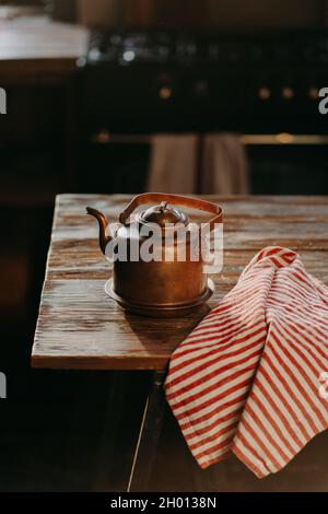 bollitore in alluminio retrò su tavolo di legno con asciugamano a righe rosso vicino. Teiera in rame usata per preparare il tè. utensili da cucina in vecchio stile Foto Stock