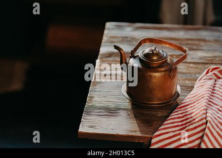 Vecchia teiera in rame metallico su tavolo in legno in camera scura. Asciugamano a righe rosso nelle vicinanze. Bollitore d'antiquariato per preparare tè o caffè. Attrezzatura di cottura Foto Stock