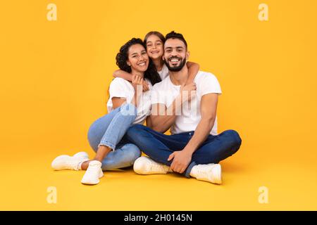 I genitori arabi e la bambina che abbraccia posando insieme seduti su sfondo giallo, sorridendo a Camera. Studio Shot della famiglia araba che abbraccia Expre Foto Stock