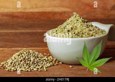 Un mucchio di semi di canapa pelati in un recipiente bianco con una lafe di canapa e un mucchio di semi di canapa non pelati davanti. Foto Stock