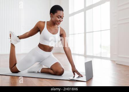 Donna africana atletica che fa esercizi di fitness sul tappeto yoga pavimento, piedi e gambe allunganti, seguendo online tutorial sport a casa. Signora che usa un computer portatile Foto Stock