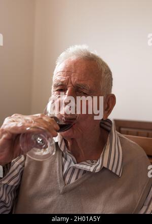 Uomo anziano in camicia e gilet bevendo vino rosso dal bicchiere a casa Foto Stock