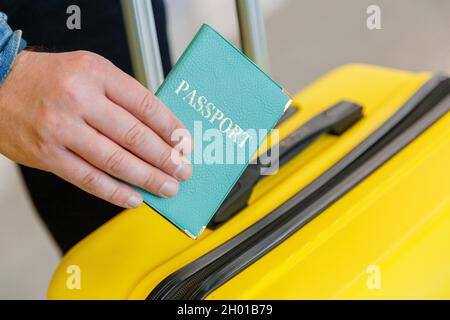 Passaporto in mano maschile dietro le valigie prima del viaggio in aereo in aeroporto. Uomo con passaporto verde e biglietto su sfondo giallo valigia Foto Stock