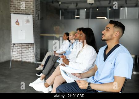 Il gruppo multirazziale di medici ha concentrato uomini e donne in abbigliamento da lavoro frequentando una conferenza medica in clinica, seduto in fila e guardando lo spazio per le copie Foto Stock