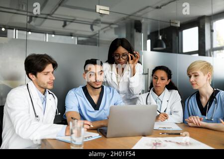 Il team professionale multirazziale di medici ha concentrato uomini e donne in workwerar seduto al tavolo, guardando lo schermo del notebook, prendendo appunti, guardando f Foto Stock