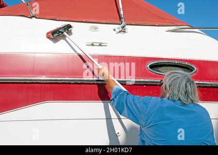Uomo caucasico anziano che lava una barca rossa e bianca con spazzola a manico lungo Foto Stock