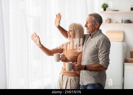 Happy Senior coppia che sventolano Ciao guardando fuori finestra tenere tazze di caffè in piedi in cucina moderna a casa. Sposi anziani in pensione gesturing Hi lo Foto Stock