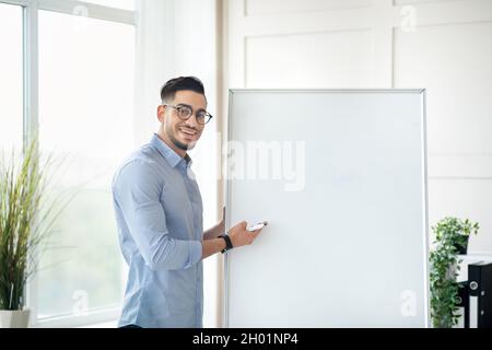 Sorridendo giovane insegnante arabo maschio in piedi vicino a lavagna vuota con mockup, dando online lezione privata da casa. Conduttore del tutor orientale millenario Foto Stock