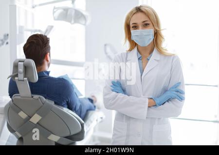 Donna dentista bionda donna in maschera protettiva del viso e guanti medici in posa in clinica dentale moderna, paziente uomo irriconoscibile seduto in ch dentale Foto Stock