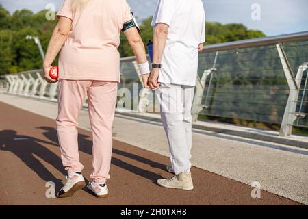 Coppia senior in tute passeggiate unire le mani lungo il ponte pedonale al tramonto Foto Stock