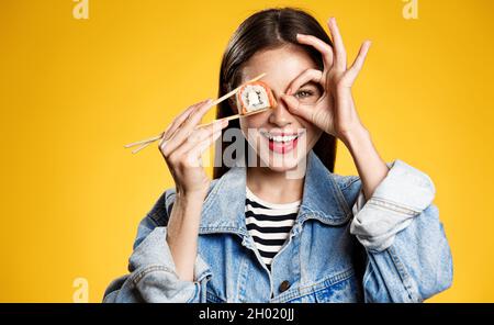 Donna sorridente che tiene sushi, sashimi con bacchette, mostrando segno giusto e sembrando felice, mangiare delicious takeaway foo, concetto di consegna di cibo Foto Stock