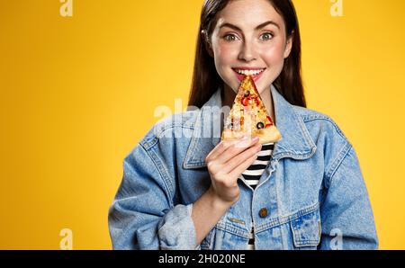 Sorridente ragazza brunetta morde la pizza, tenendo fetta nei denti e guardando felice, concetto di fast food consegna e mangiare fuori, sfondo giallo Foto Stock
