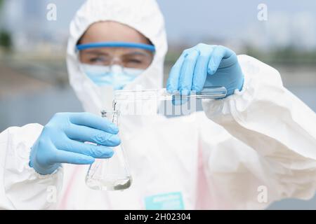 Lo scienziato in tuta protettiva con bicchieri versa l'acqua dalla provetta al pallone Foto Stock