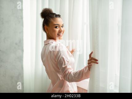 Nuovo giorno di benvenuto. La donna allegra è svegliata e sta in piedi vicino alla finestra, aprendo le tende e sorridendo alla macchina fotografica. Giovane bella donna in pigiama svegliarsi Foto Stock