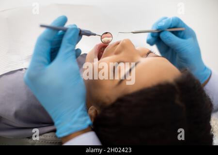 Giovane donna nera seduta in sedia durante il trattamento dentale con stomatologo, medico dentista irriconoscibile in Blue Guanti sterili tenuta troppo Foto Stock
