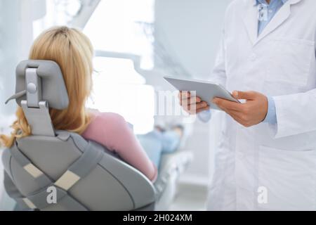 Tecnologie moderne e concetto di stomatologia. Corto di dentista maschile in possesso di nuovo tablet digitale su una donna bionda irriconoscibile seduta paziente Foto Stock