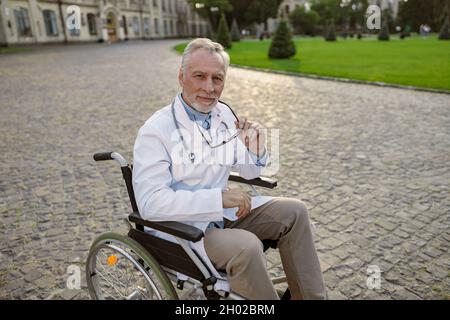 Fidato medico senior handicappato in sedia a rotelle indossare camice da laboratorio e occhiali guardando la macchina fotografica, in posa all'aperto Foto Stock