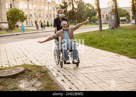 Eccitato uomo maturo disabili in sedia a rotelle indossando cuffie divertirsi durante una passeggiata in città assistita da giovane infermiere in maschera protettiva viso Foto Stock