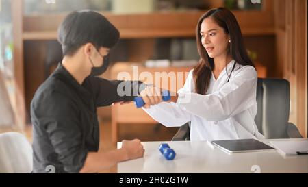 Una terapeuta femminile asiatica aiuta un paziente maschio ad allungare la sua parte superiore del braccio utilizzando un piccolo manubri dopo l'intervento chirurgico. Centro clinico di terapia Foto Stock