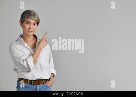 Donna asiatica di mezza età che punta le dita verso l'alto nello spazio vuoto per il testo isolato su sfondo bianco. Guardando la fotocamera Foto Stock