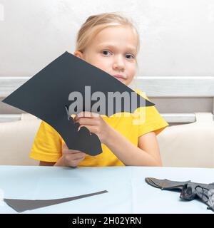 Una ragazza taglia una decorazione di Halloween a casa da carta nera. Fai da te artigianato decorazioni per la casa. Il bambino fa i mestieri di carta, origami Foto Stock