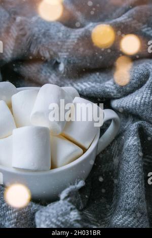 Marshmallows bianchi in una tazza bianca su sfondo maglione. Calda bevanda invernale al cioccolato cacao. Luci di Natale. Preparazione accogliente per l'atmosfera di vacanza, Foto Stock